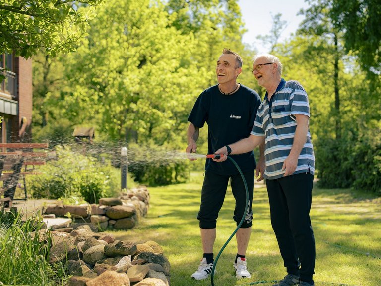 Zwei Männer stehen mit einem Gartenschlauch in der Hand vor einem Blumenbeet und gießen die Pflanzen. Im Hintergrund sind grüne Bäume und Sträucher zu sehen.