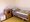 Ein einfaches Pflegezimmer mit einem Einzelbett, bezogen mit grau-grün gestreifter Bettwäsche. Daneben steht ein hölzerner Nachttisch mit einem Telefon und ein kleiner weißer Nachtschrank auf Rollen. Der Raum verfügt über weiße Wände und einen gelben Boden.
